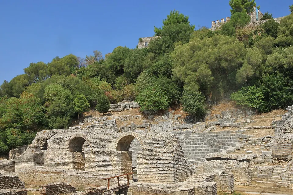 The stone tiers and arched stage building of an ancient theatre set into a wooded hillside, with the walls of a castle just visible at the top of the hill.