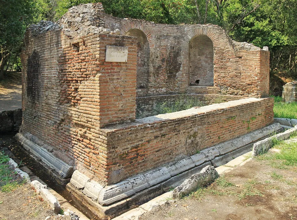 A rectangular fountain house of red brick and stone with two arched niches in its back wall and a basin in front, on a grassy site among trees.