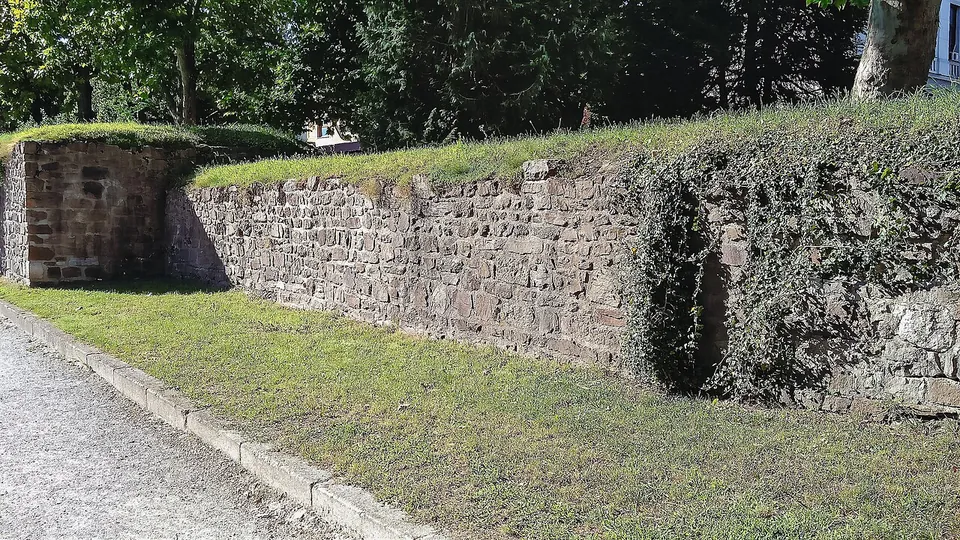 Close view along the wall's face of irregular brown and grey stones laid in rough courses, partly overgrown with ivy, with a square tower at the left and grass along the top.