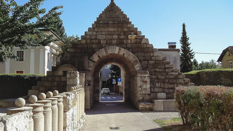 A round-arched passage through the wall under a tall stepped pyramid of squared stone blocks, with a balustrade of stone balls in the foreground and a street visible through the arch.