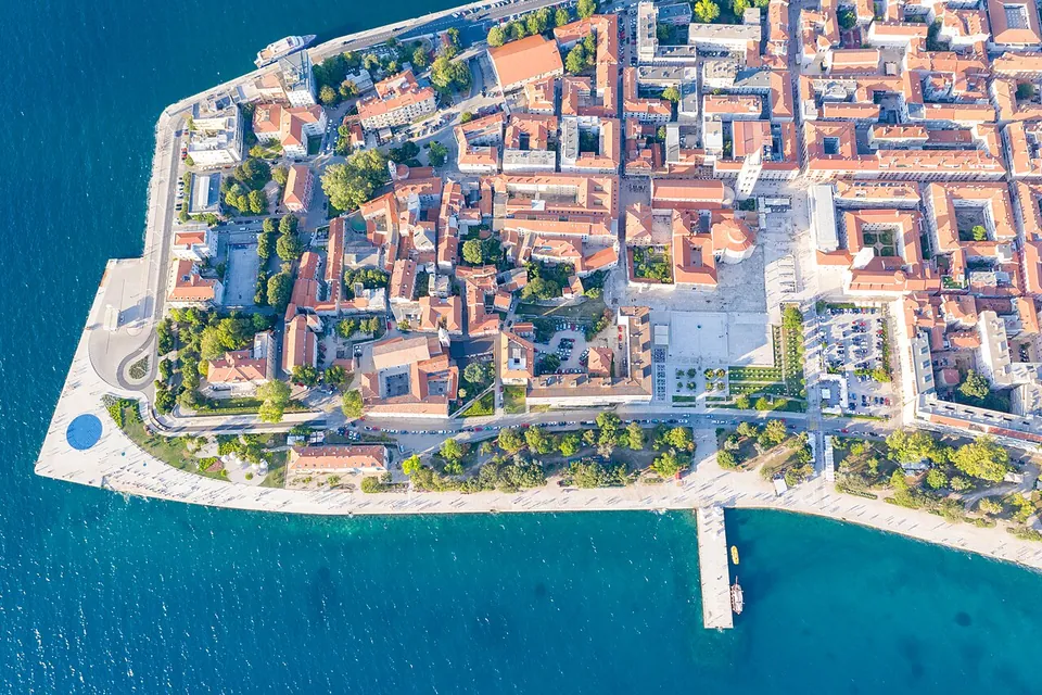 An aerial view of a narrow peninsula packed with red-roofed old town buildings, edged by a seafront promenade with a pier, with a large open square visible among the roofs.