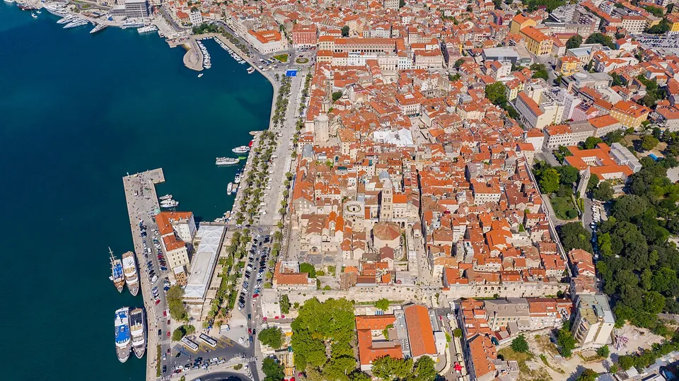 An aerial view of a dense old town of pale stone buildings with red-tiled roofs beside a harbour of blue water, a palm-lined promenade along the waterfront and moored boats at a pier.