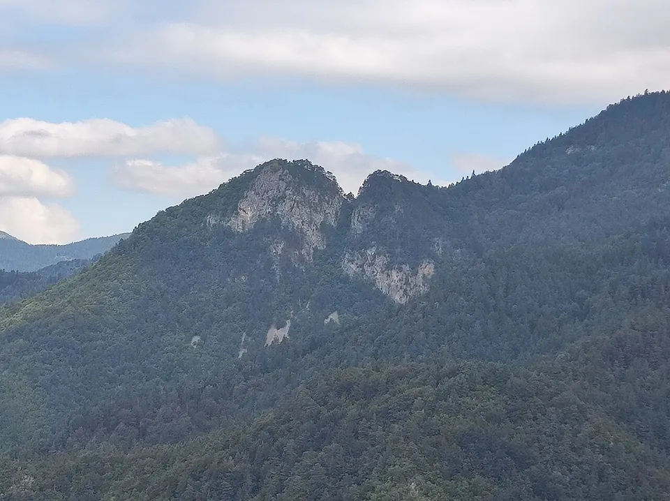 A forested mountainside with two pale rocky crags standing out near its top, under a cloudy sky.