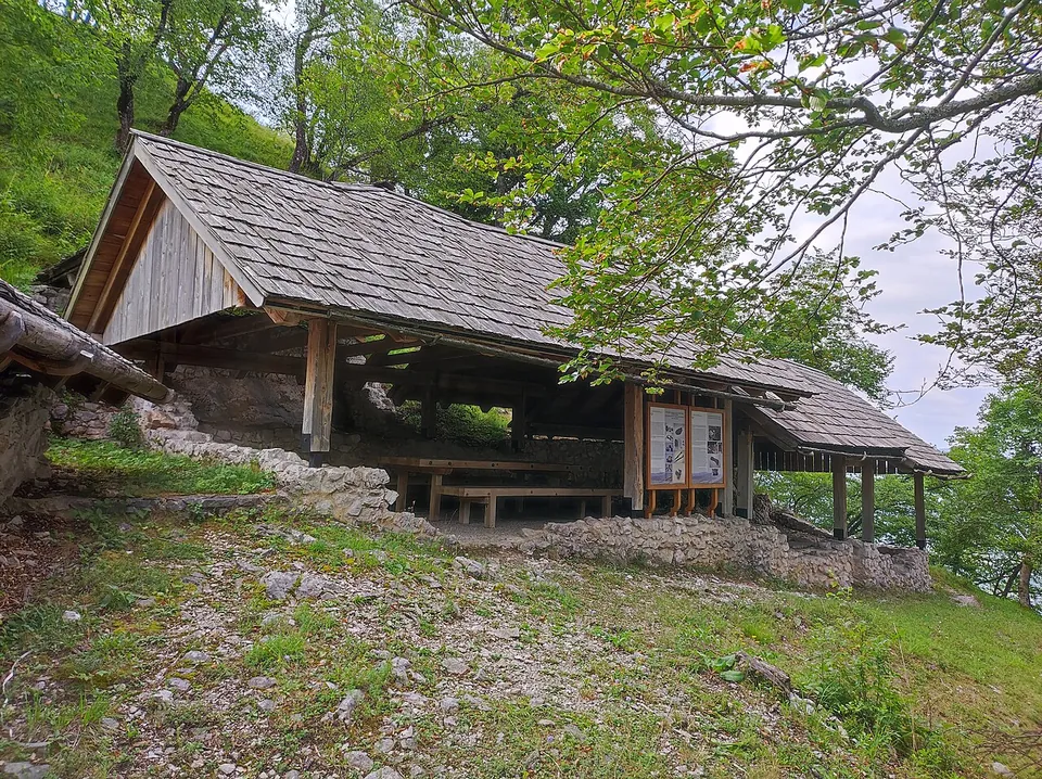 A timber shelter with a shingled roof built over stone wall footings on a slope, with a bench beneath it and an information panel at one end.