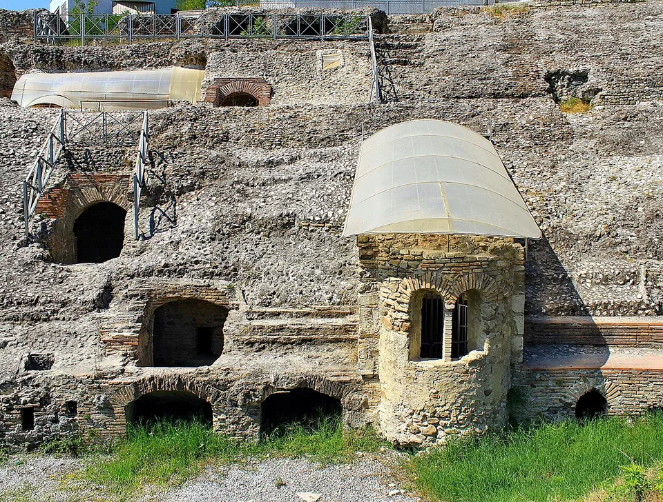A small round stone chapel with narrow arched windows under a translucent protective roof, built against the terraced ruins of the amphitheatre.