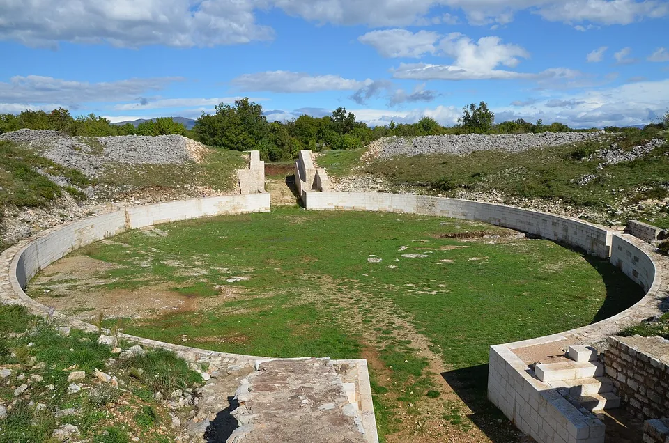 The oval grassy arena of an amphitheatre enclosed by a low curved white wall, with a narrow entrance passage in the foreground and the rubble slopes of the seating behind.