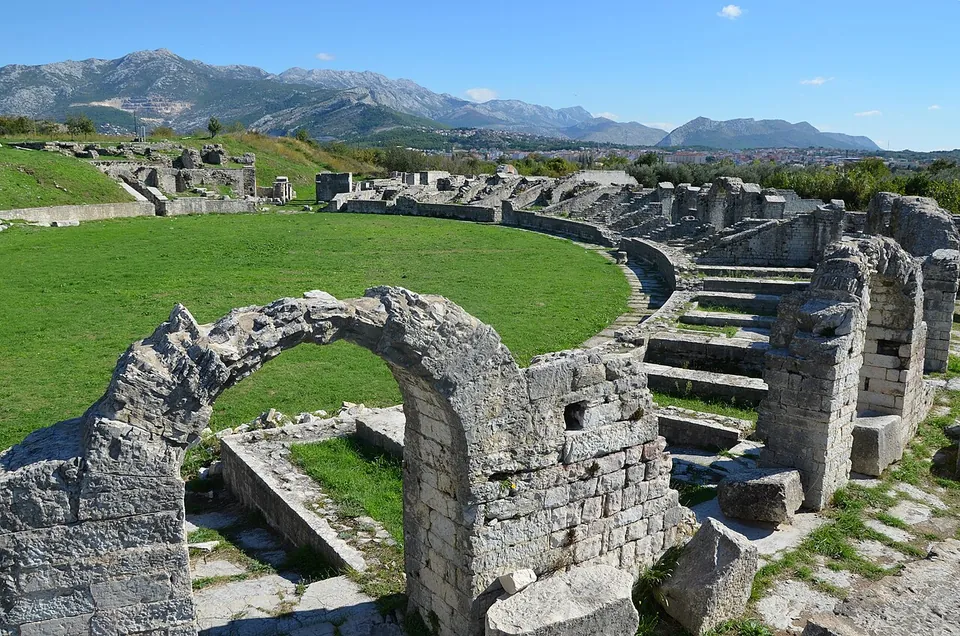 The oval of a Roman amphitheatre with a lawn for its floor, surrounded by the broken lower walls and stairs of its seating, a single stone arch standing in the foreground and mountains beyond.