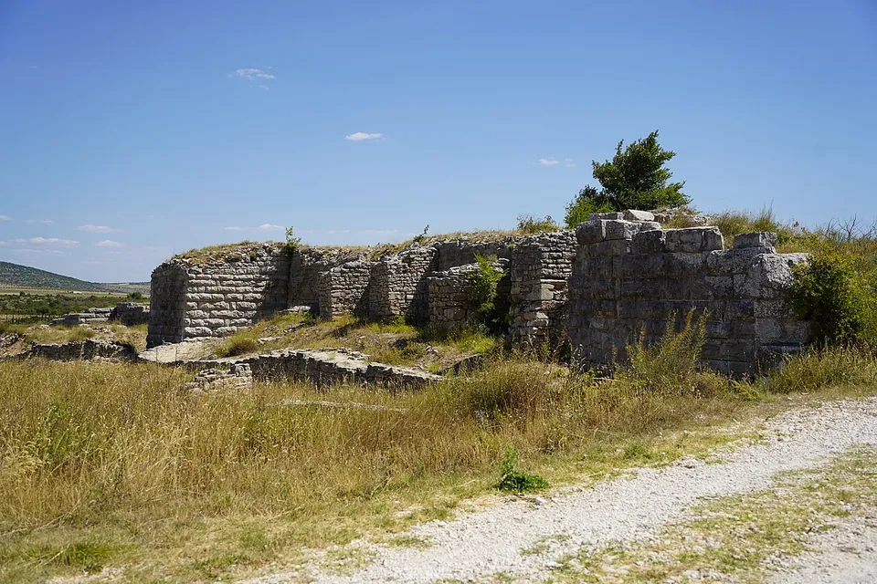 Ruined walls of large squared stone blocks with a projecting tower standing on a rise above dry grass and scrub, with a gravel track leading towards them under a blue sky.