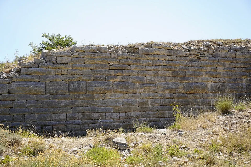 A long stretch of wall built of horizontal courses of pale squared limestone blocks, with dry grass growing along its top and at its foot.