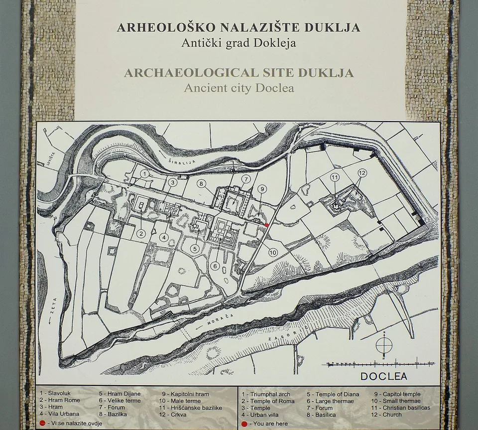 An information board headed "Arheološko nalazište Duklja / Archaeological site Duklja", with a drawn plan of the walled town between two rivers and a numbered key to its buildings in Montenegrin and English.