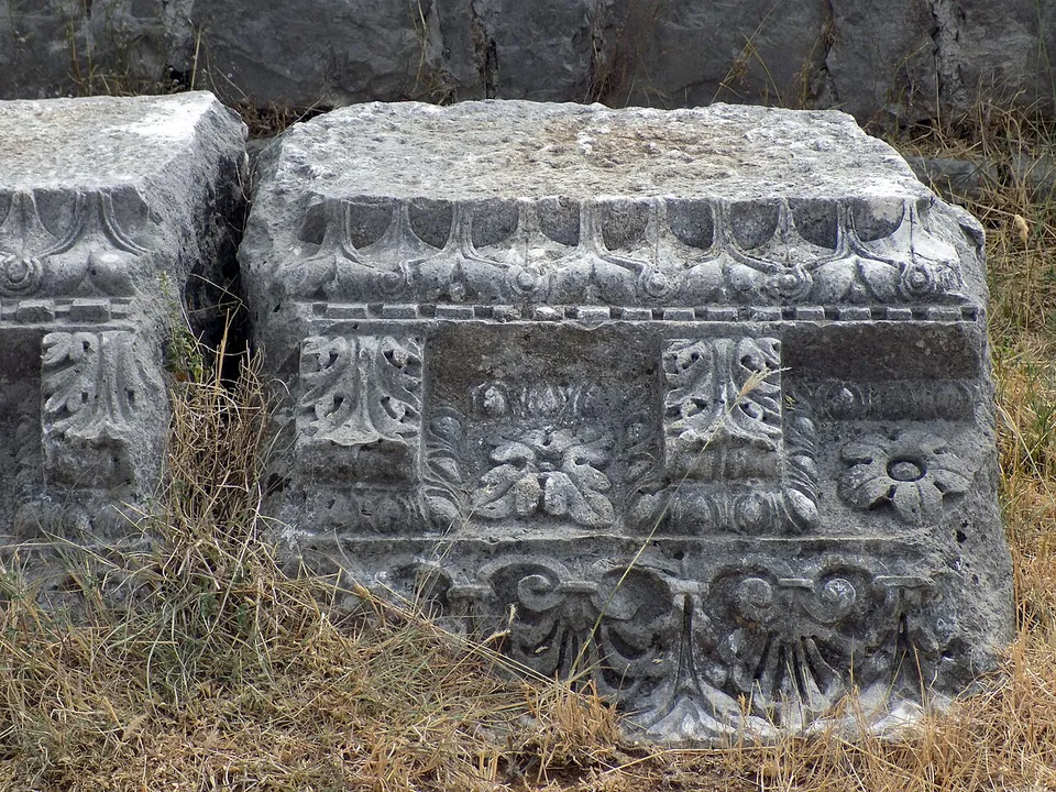 Close view of weathered limestone blocks lying in dry grass, carved with egg-and-dart mouldings, acanthus leaves and rosettes.