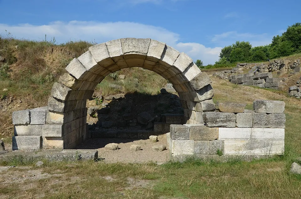 A single round arch of large squared stone blocks framing a dark opening into a hillside, with dry grass around it.