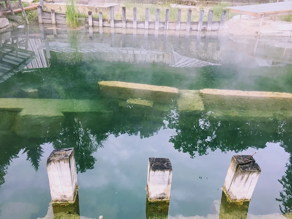 Mist rising from still green water in a stone-lined basin, with the stumps of three square stone pillars in the foreground and the rim of the basin just under the surface.