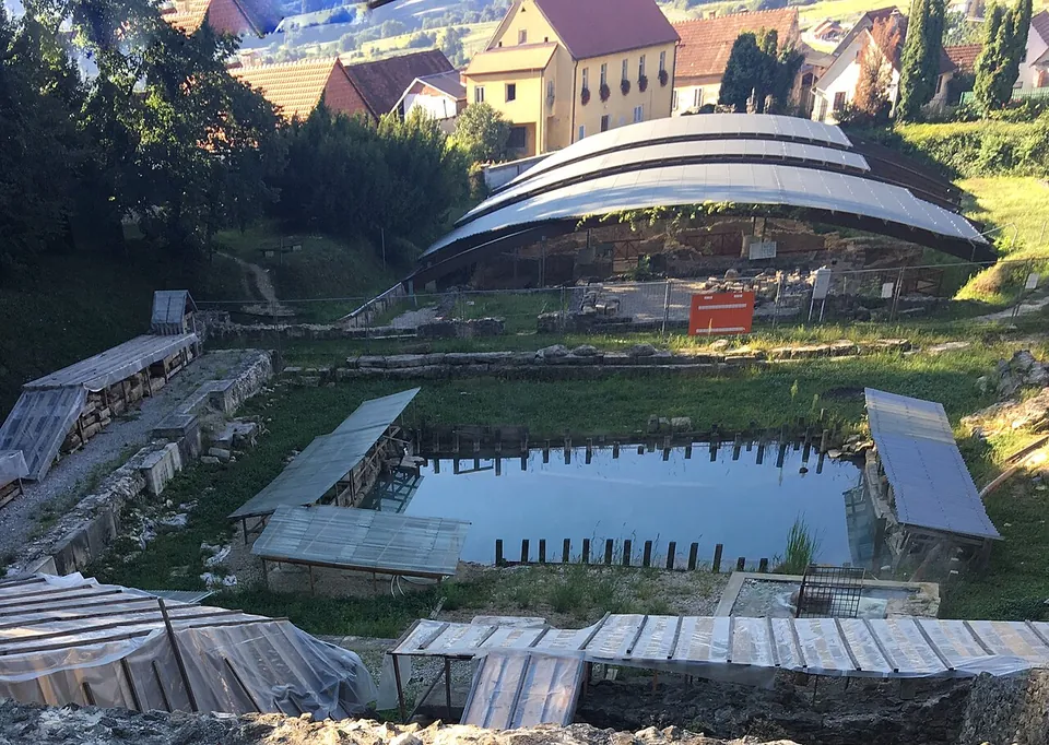 Looking down on an archaeological park of low Roman walls under long curved metal roofs and plastic covers, with a rectangular pool of water in the middle and the houses of a small town behind.