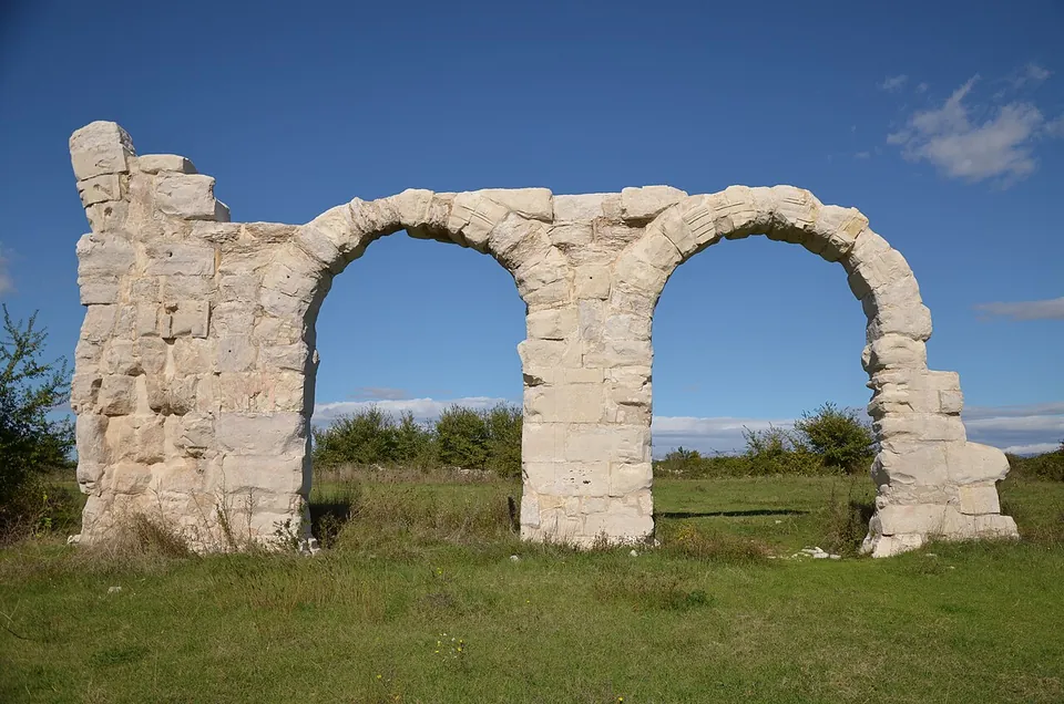 Two round arches of large pale stone blocks on three piers, the end piers broken off at different heights, standing on grass against a blue sky.