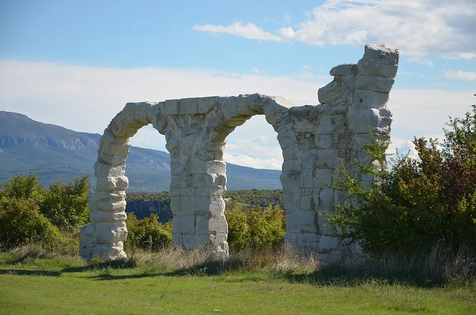 Two tall round arches of pale limestone blocks and a broken pier standing alone in a meadow, with scrub around them and a long mountain ridge beyond.