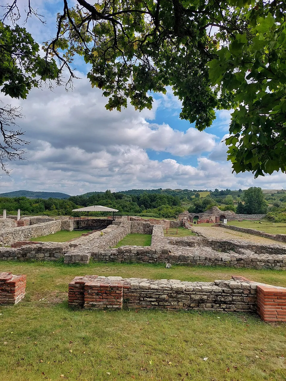 Low stone and brick foundation walls spread across mown grass inside the enclosure, a white canopy covering one excavated area, with wooded hills on the horizon.