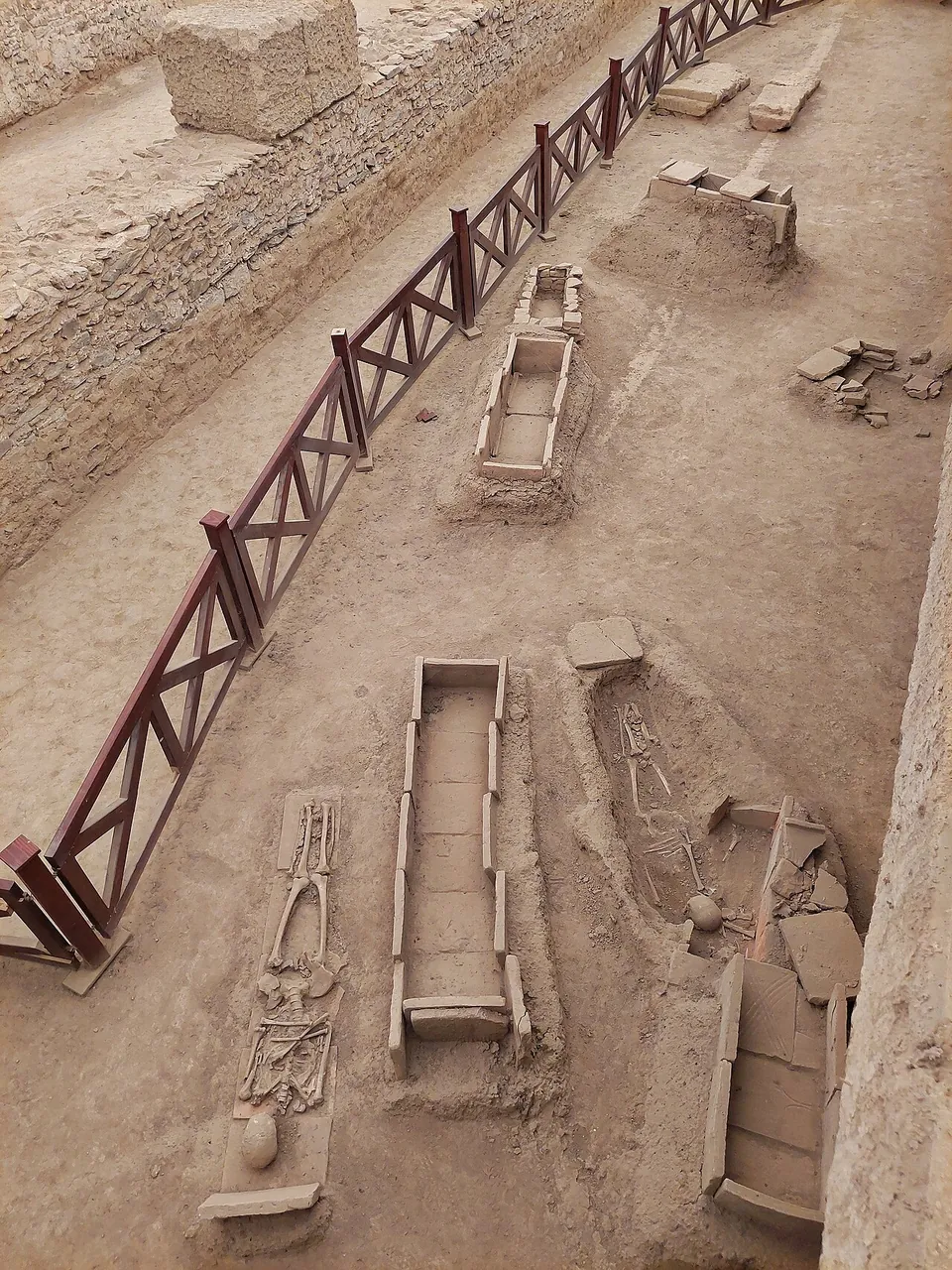 Excavated graves seen from above, some stone-lined and empty, others holding skeletons still in place, with broken tiles and a wooden handrail along the edge of the cutting.