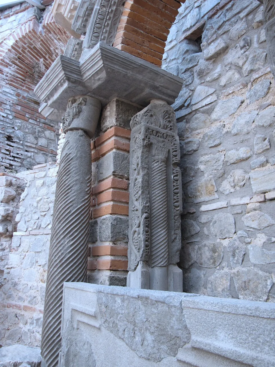 A spiral-fluted stone column beside a pilaster carved with dense relief ornament, together carrying a moulded cornice, set into a wall of rubble stone banded with red brick.