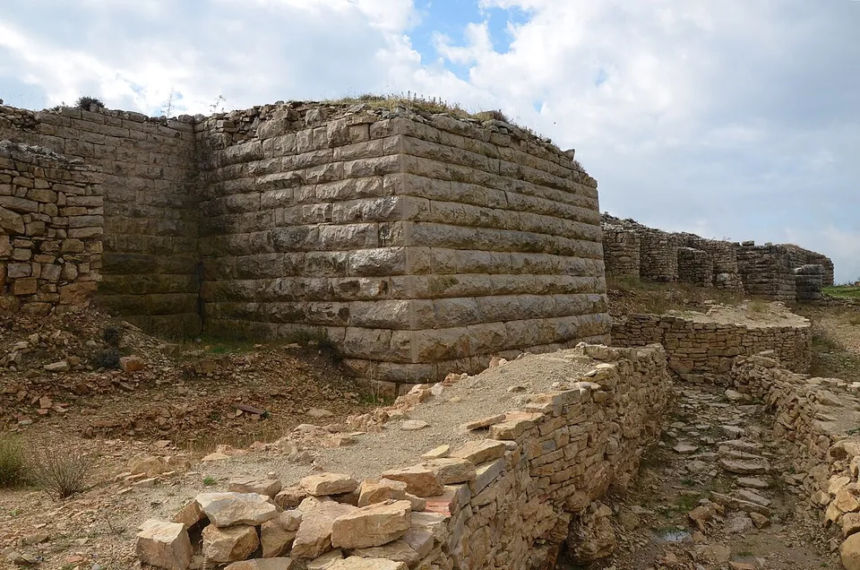 A corner of massive city wall built of long courses of squared limestone blocks, standing several metres high, with lower stone foundations and a rubble path in front of it.