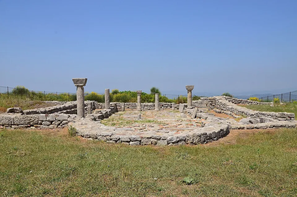Low stone walls and fallen blocks of a church with a few column shafts standing, one with its capital, on a hilltop with hazy mountains beyond.