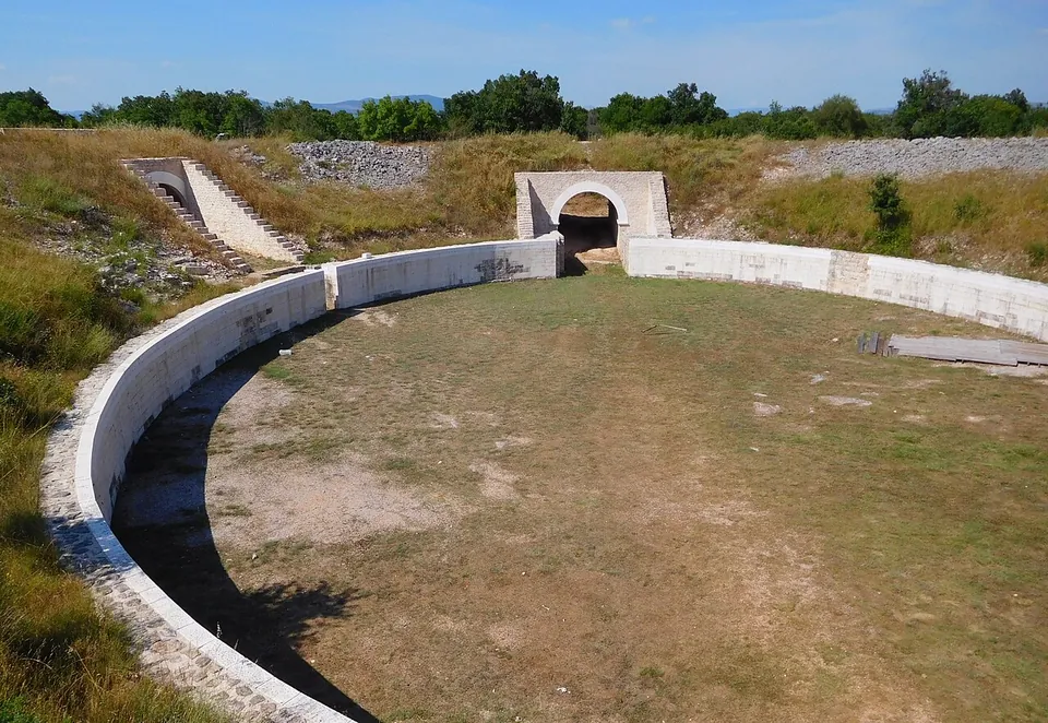 A passage between two walls of neatly coursed pale limestone blocks, their ends stepped, leading into the grassy oval of an amphitheatre.