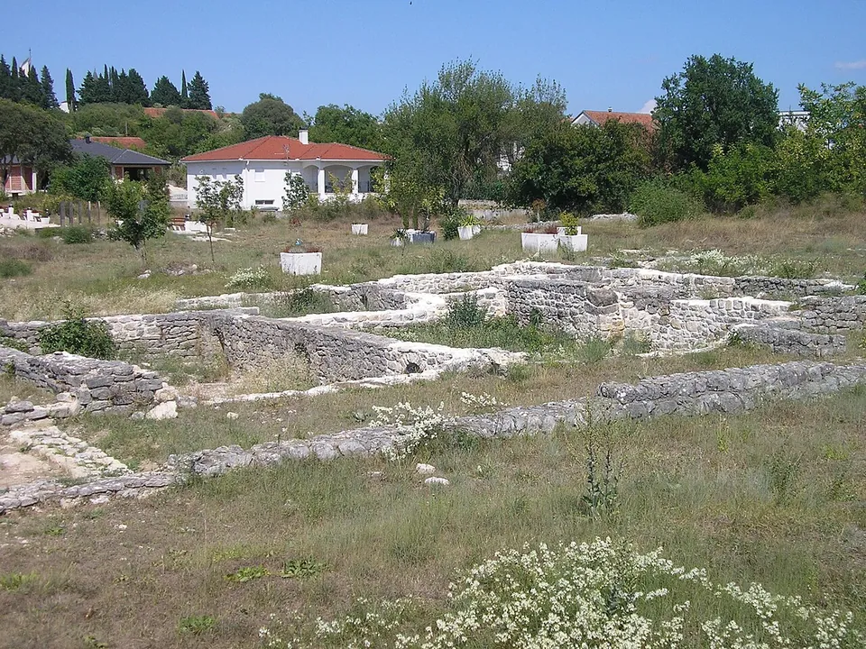 Low conserved stone wall footings of several rooms spread across a dry meadow, with small white stone markers, trees and a red-roofed house behind.