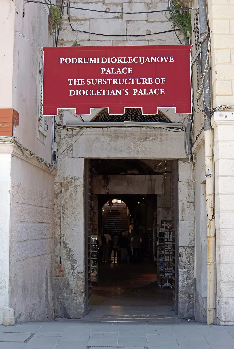 A narrow stone doorway in a pale wall under a red sign reading "Podrumi Dioklecijanove palače / The substructure of Diocletian's Palace", with a vaulted passage beyond.