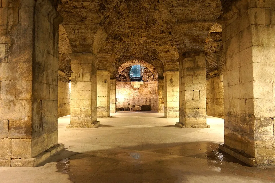 A long underground hall of massive limestone piers carrying rough vaults, lit warmly from below, with a patch of wet floor in the foreground and a lit window at the far end.