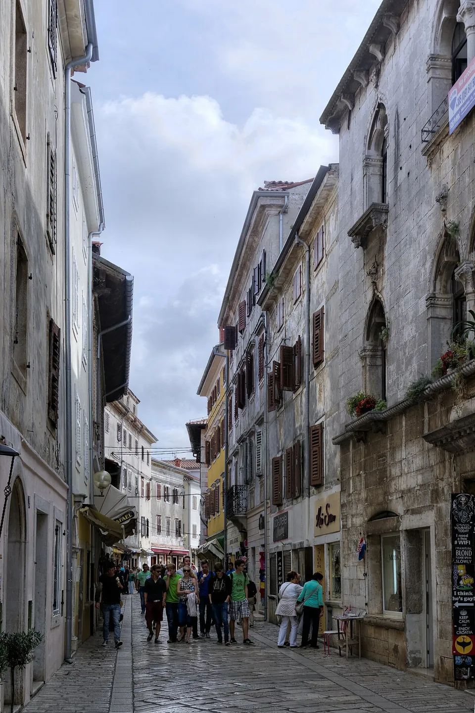 A narrow paved street between tall stone town houses with balconies and shop signs, busy with people, running straight into the distance under a cloudy sky.