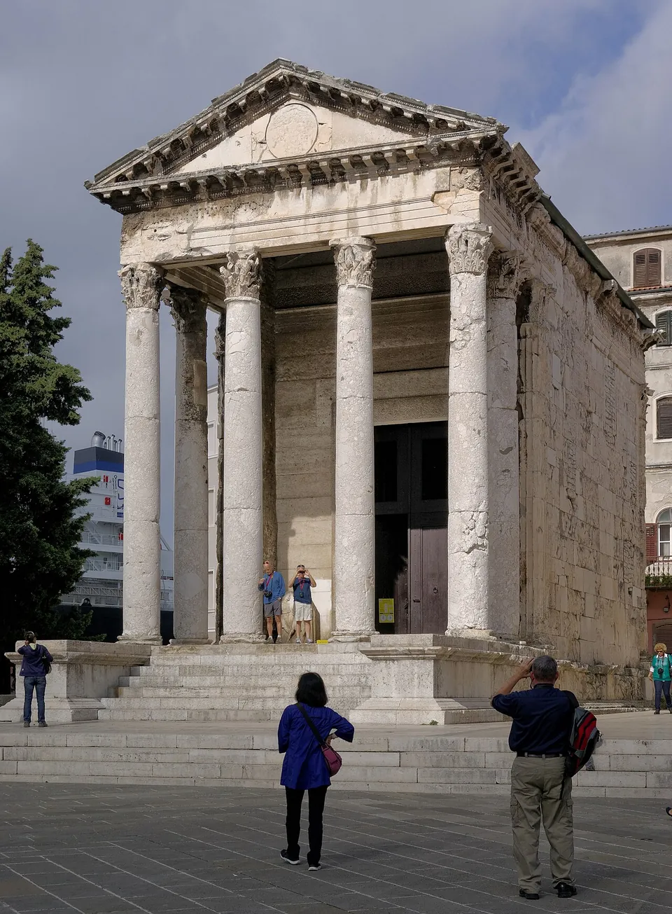 A small Roman temple on a high podium with a flight of steps, its porch of tall Corinthian columns carrying a carved frieze and a triangular pediment, with visitors standing on the square in front.