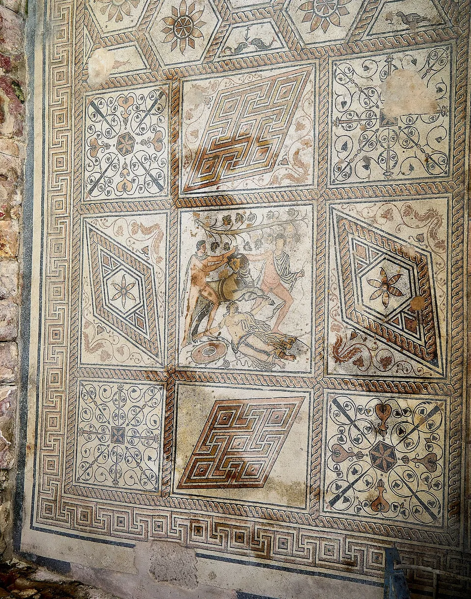A Roman floor mosaic of geometric panels, lozenges, stars and meanders in red, black and white, with a central figured panel showing a struggle between figures and a bull.