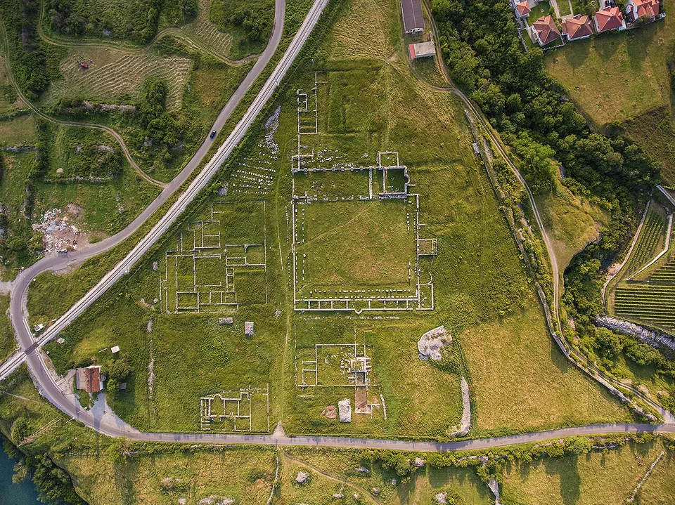 An aerial view of a long green plateau between roads and a river, marked with the rectangular outlines of excavated Roman buildings, a large square courtyard among them, with houses and fields around.