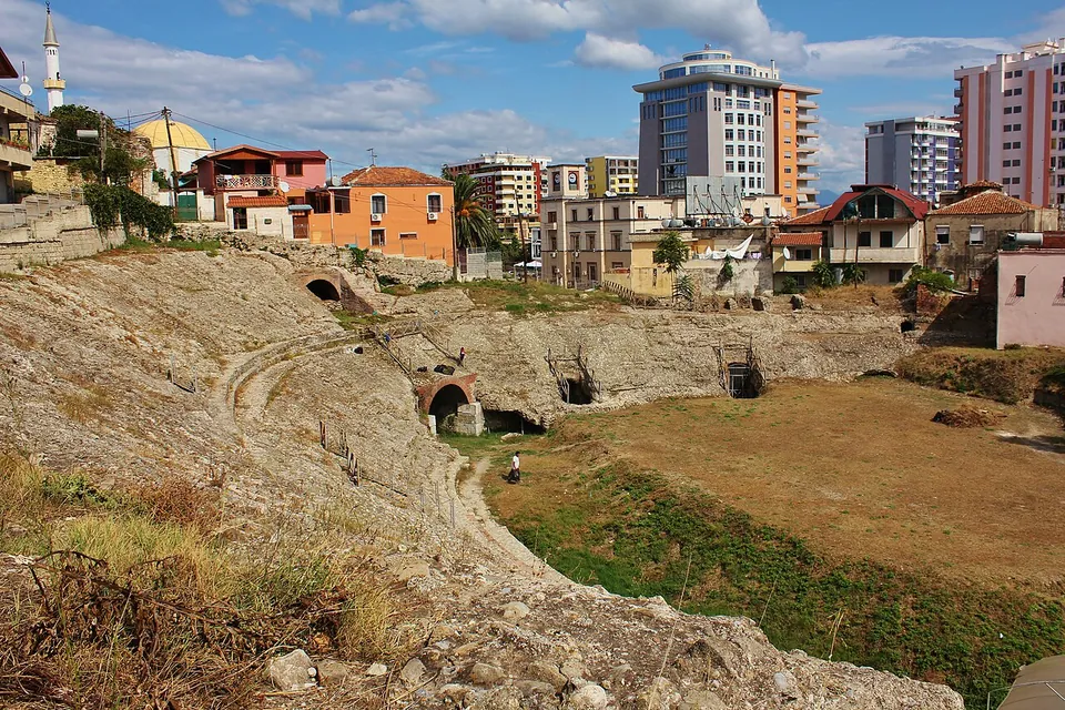 The curved slope of a ruined amphitheatre's seating with brick-arched openings, surrounding a dusty arena, with houses and modern apartment towers crowding round it.