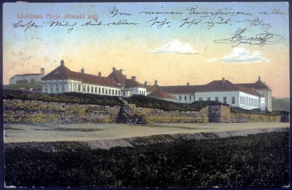 A hand-coloured postcard with a handwritten message across the sky, showing a long, low wall with grassy slopes in front of a row of large red-roofed buildings.
