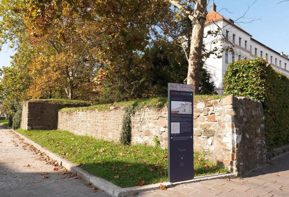 A stretch of rough stone wall capped with turf, with a square tower projecting at its far end, a paved pavement in front and a dark information stele standing against it; autumn trees and a large white building behind.