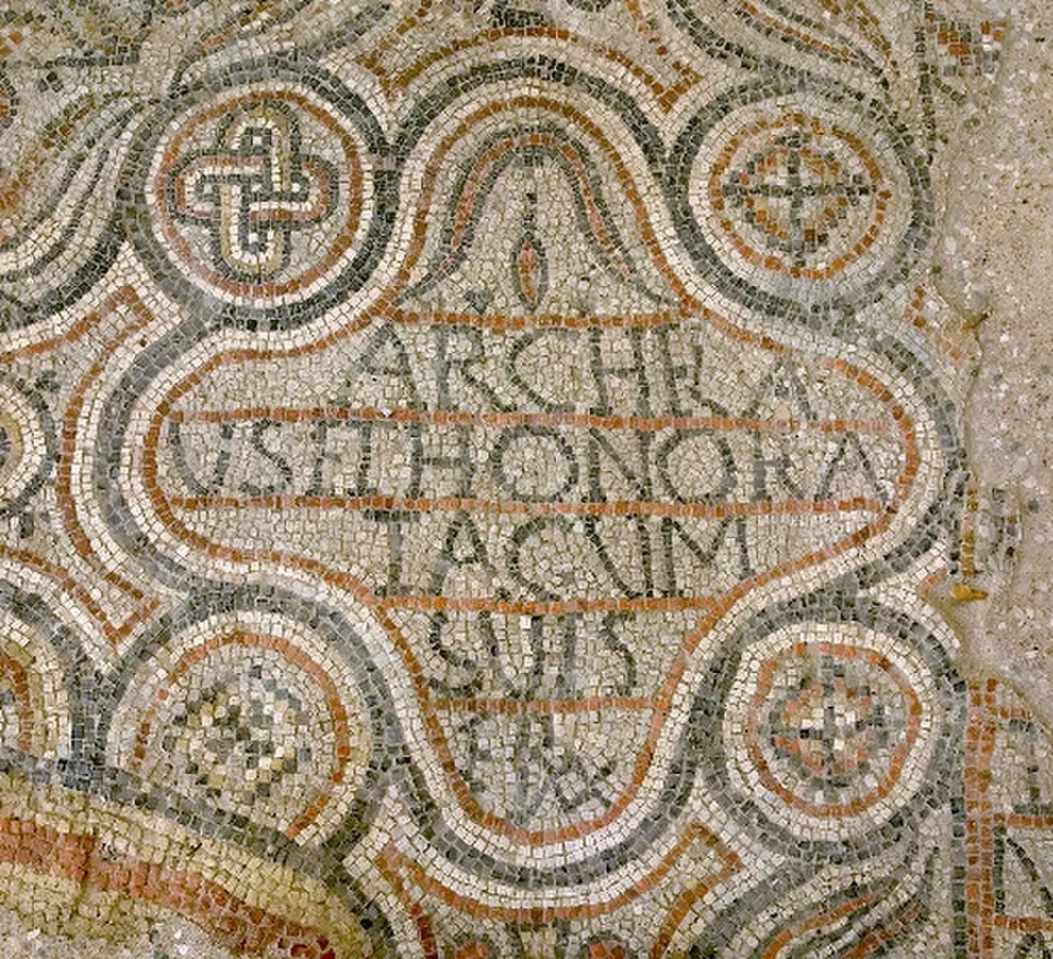A mosaic panel of small white, grey and red stones, an inscription in capital letters framed by a looping border with cross motifs in roundels around it.