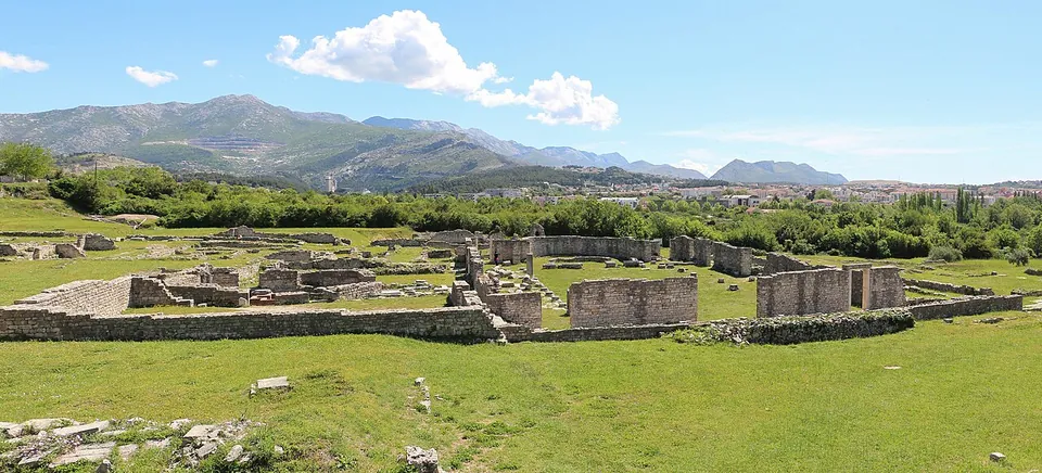 A wide green field scattered with the low stone walls and foundations of Roman buildings, with trees behind and bare limestone mountains on the horizon under a blue sky.