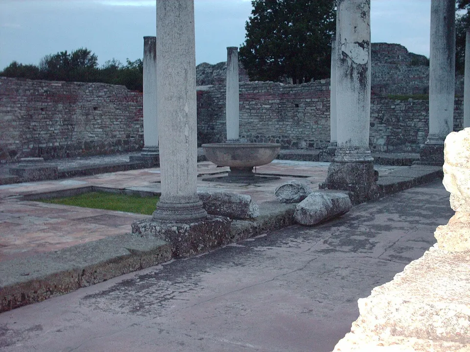Columns standing on a paved floor around a shallow stone basin, with excavated walls and trees behind them in evening light.