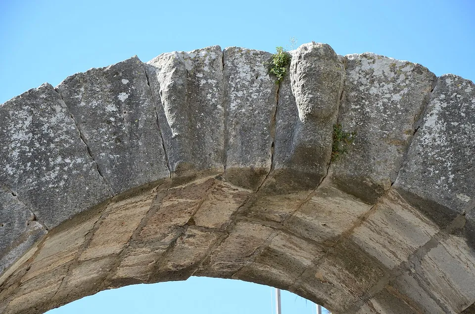 Close view of a weathered stone arch against a blue sky, with a worn relief of a bearded head at the crown of the arch and a small plant growing from a crack.