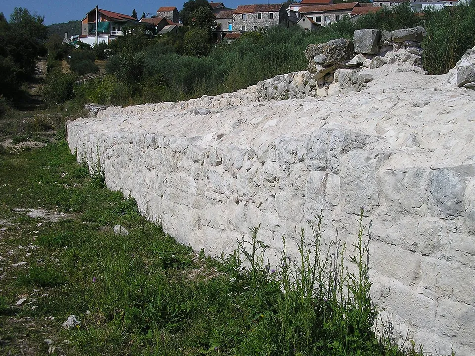 A long, low wall of large pale stone blocks along a grassy slope, with houses of the village above.
