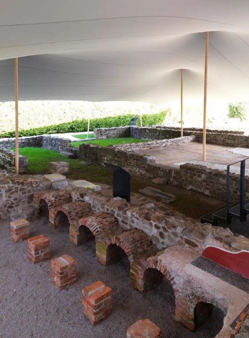 A row of small round-headed brick arches and free-standing square brick stacks set low in the ground under a white fabric canopy, with stone wall footings and a lawn beyond.