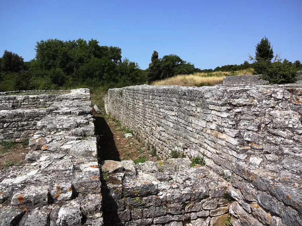 Two long parallel walls of roughly coursed grey stone about head high, with a narrow gap between them, rising from dry grass with trees behind.