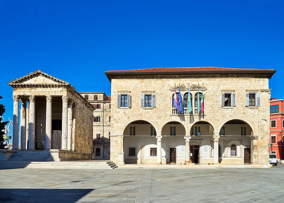 A paved square under a deep blue sky with a small classical temple on a high podium on the left and a pale stone Gothic town hall with a ground-floor arcade and flags on the right.