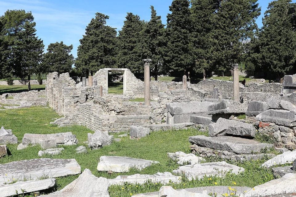Tumbled stone blocks and slabs in the grass in front of the low walls of an early Christian basilica, with two slender columns standing and a line of dark cypresses behind.