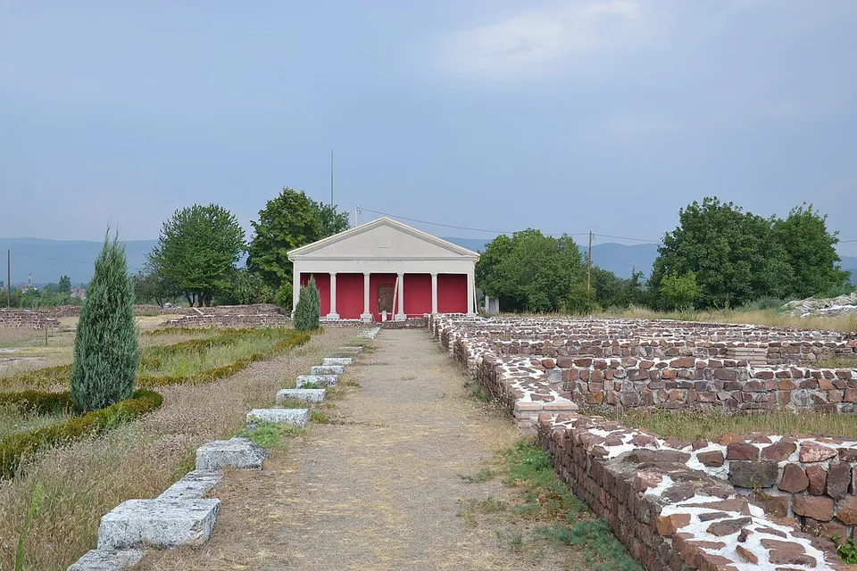 A gravel path running between low rubble wall foundations and a line of stone blocks, towards a small modern pavilion with a triangular pediment and red panels between white columns, with hills on the horizon.