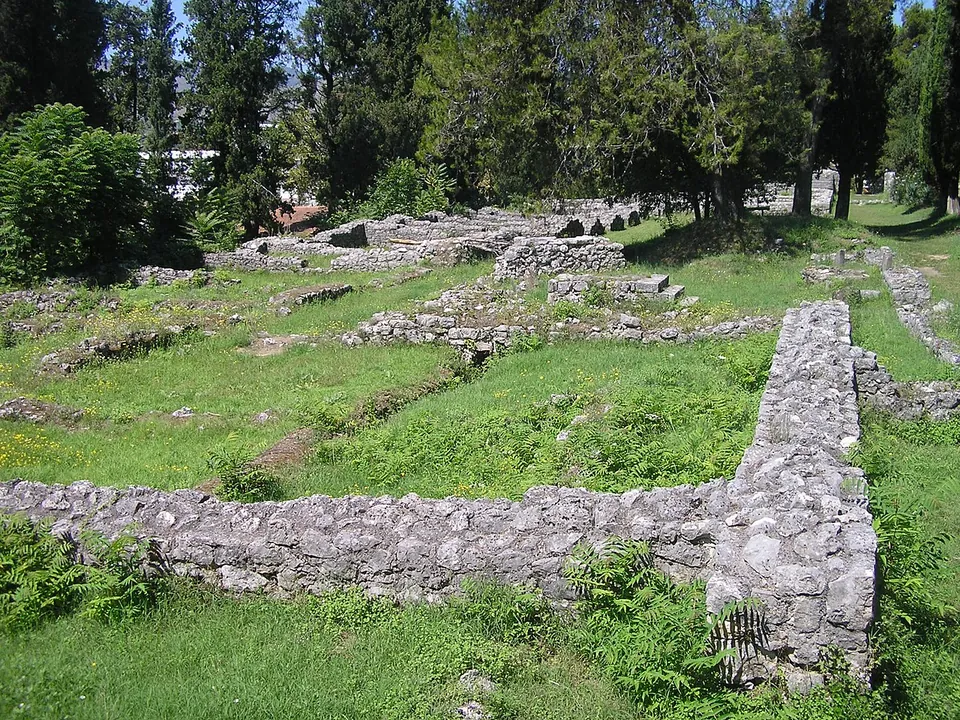 Long low walls of grey rubble stone crossing a lawn in a grove of dark cypress trees, outlining rooms of a large building.