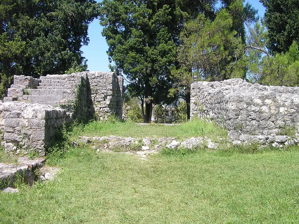 A gap in thick stone walls with steps rising on the left and a low sill across the grassy opening, under cypress and pine trees.