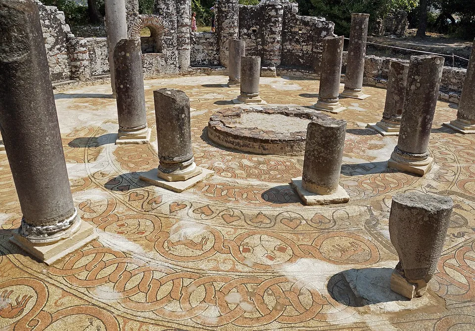A round mosaic floor of interlaced roundels in red, ochre and cream, with a ring of granite column stumps and a circular basin at the centre, under a clear sky.