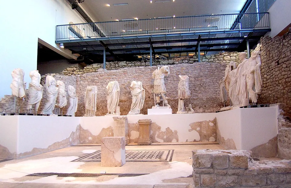 A row of headless and damaged white marble statues of men and women in togas and robes standing on a plinth along the back of an excavated temple room with a mosaic floor, under a modern metal gallery.
